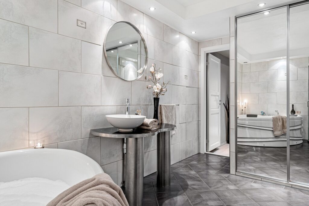 A grey-panelled ensuite bathroom with a grey-tiled floor, chrome fixtures, and a circular mirror on the wall.