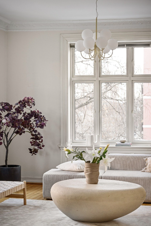 White living room with light wood flooring and a white rug, with a stone coffee table and purple potted tree