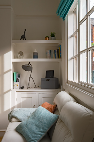 White sofa with blue and orange cushions underneath a window, next to white shelving with books and ornaments