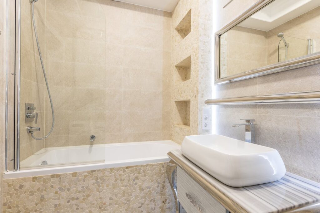 A bathroom with a glass-screened bath, beige tiling of various shapes and sizes, and an unorthodox-shaped basin.
