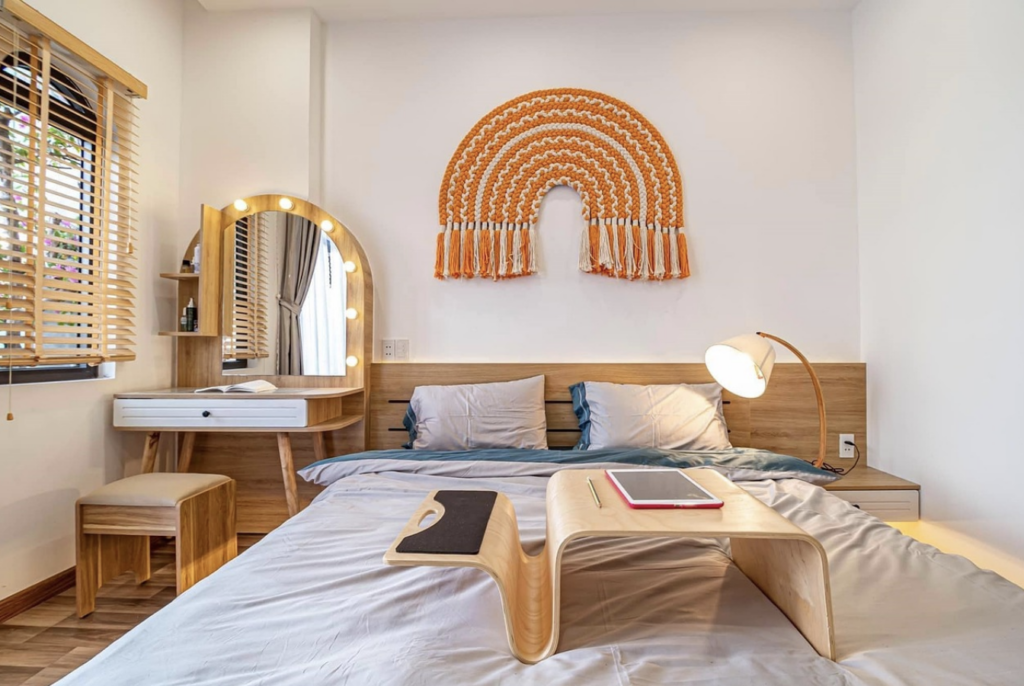 a modern, white bedroom featuring a light-coloured bed with a wooden half-panelled wall, a wooden nightstand with a lamp, a mirrored vanity, and an orange rainbow hanging decoration above the bed.
