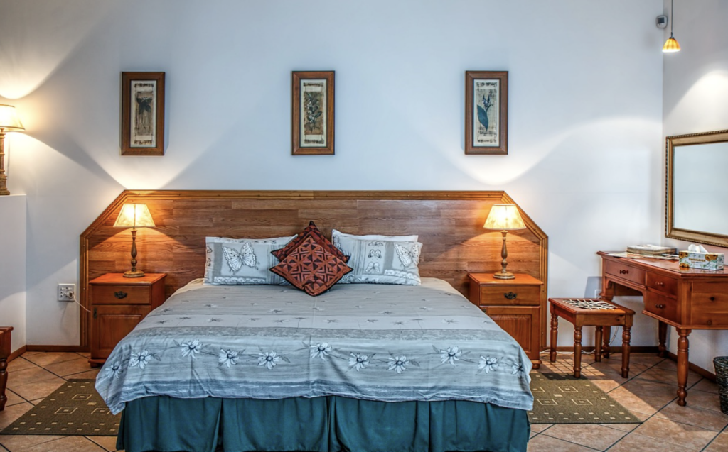 a neutral-toned bedroom with a dark wooden panelled headboard and matching wooden bedside tables, a blue patterned bed, and beige tiled flooring.