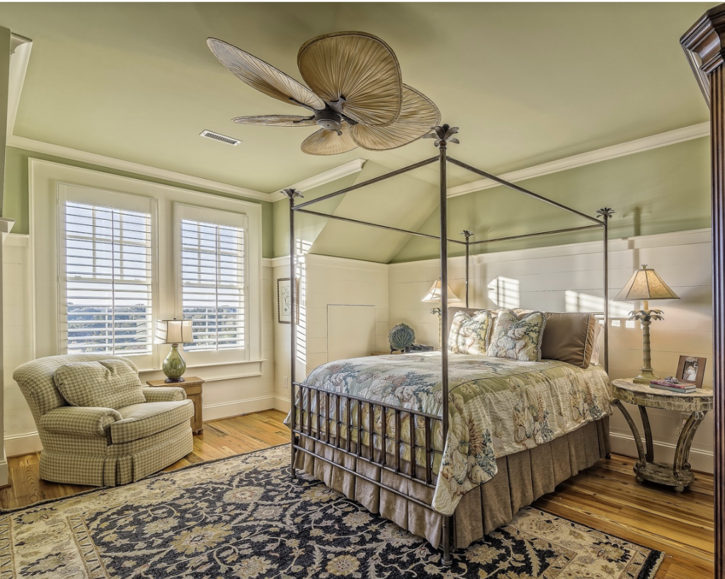 a light green bedroom featuring white half-wall panelled sections and a navy patterned rug, with a four-poster bed and green armchair.