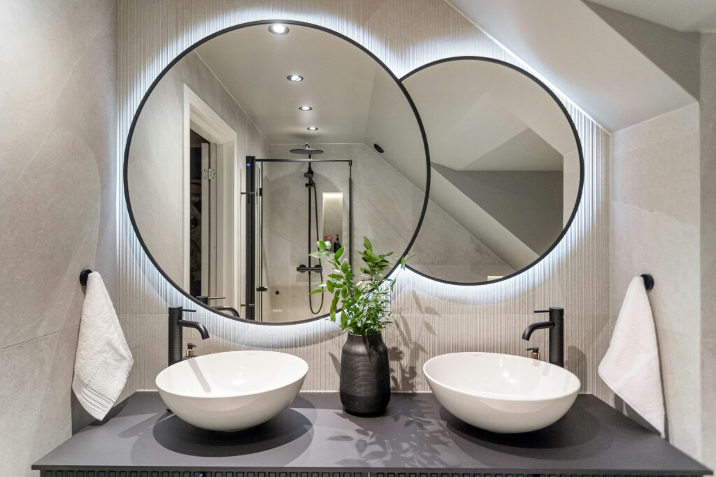 two black-framed circular mirrors lit by LEDs hung above two white bathroom sinks with contrasting black taps and a black worktop