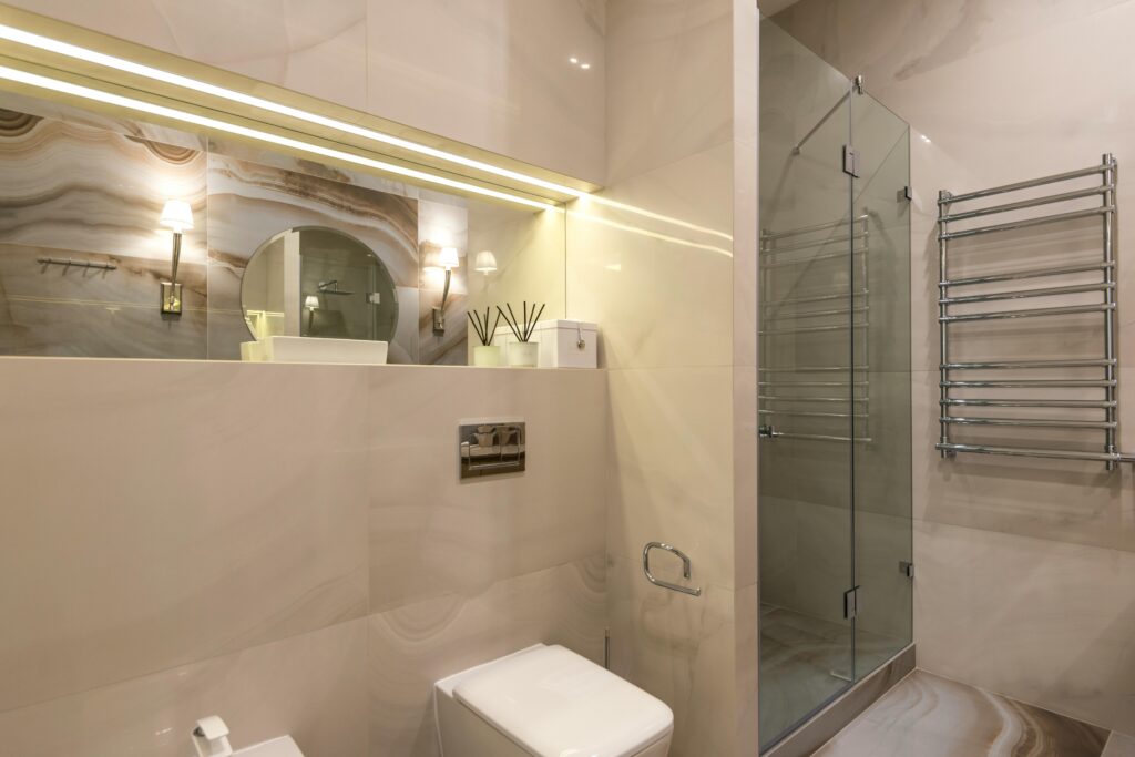 A neutral-toned bathroom with an LED-lit shelf, a walk-in shower, and a chrome towel rail on the wall