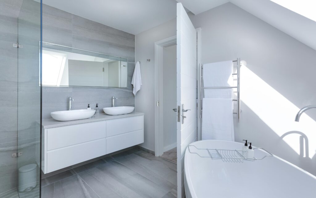 A light grey and white bathroom with two sinks, a large rectangular mirror, and a white bathtub in the corner