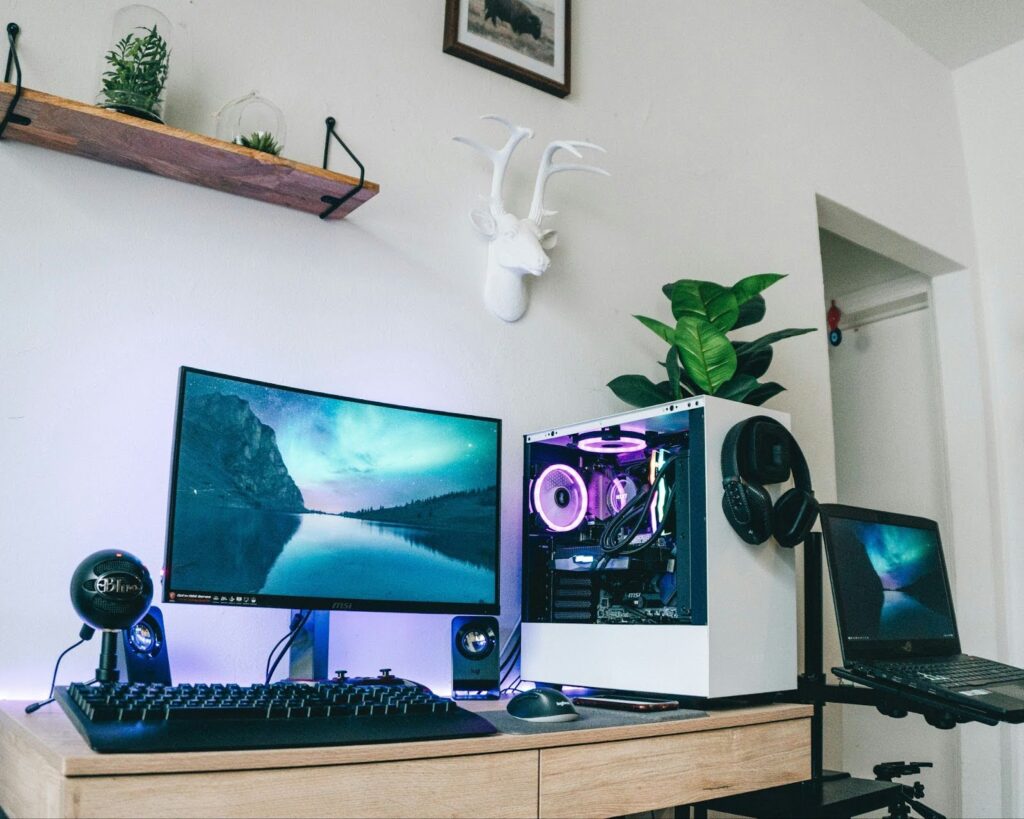 A gaming room with white walls, a wooden desk setup with a monitor, PC, and a headset.