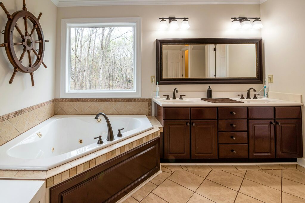 A beige tiled bathroom with a white corner bath, dark wooden cabinets and a wooden ship wheel hung on the wall.