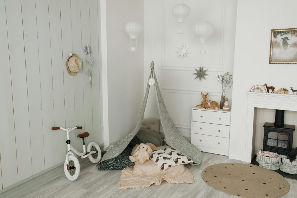 Neutral toned children’s bedroom with a white wooden wall panel accent wall, white walls and grey wood-effect floors. It’s decorated with animal toys, a grey pitch tent, bicycle and round beige carpet.