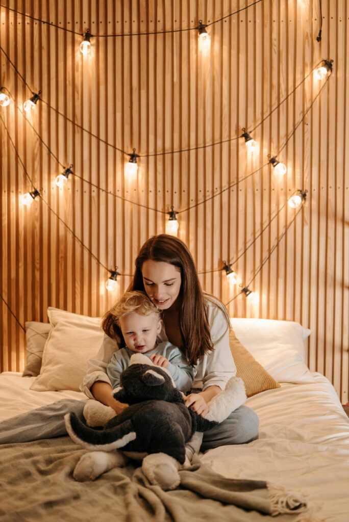 A child's bedroom that is fitted with acoustic slat wall panels, a white bed and warm fairy lights. On the bed a woman and child are playing with a stuffed animal.