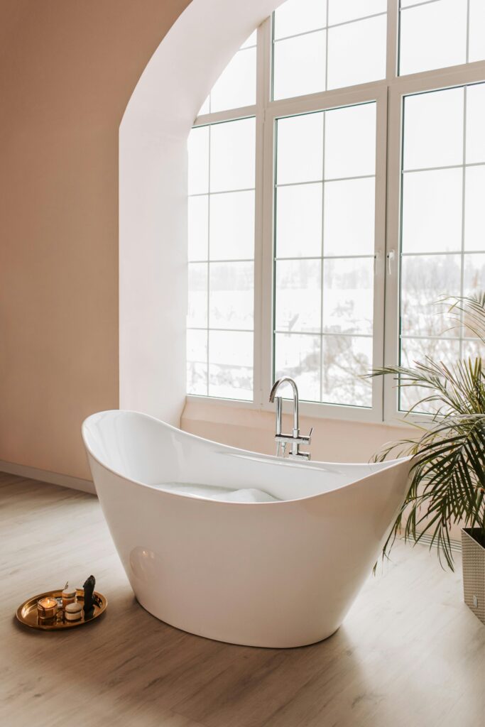A white freestanding bath in the centre of a beige, minimalistic bathroom and in front of a large, panelled window.